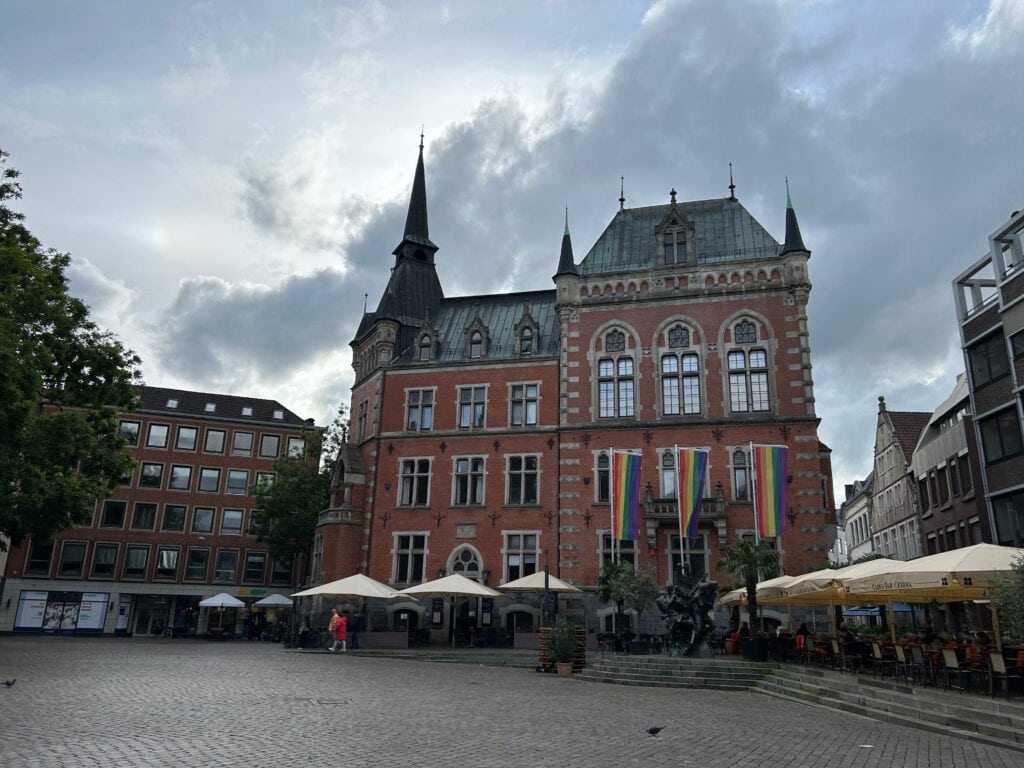Historisches Gebäude mit Regenbogenfahnen und Cafés, bewölkter Himmel.
